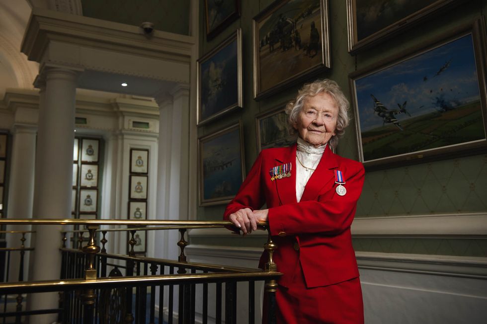 Dorothea Barron dressed in a red suit with medals on her front, standing in from of paintings of wartime scenes