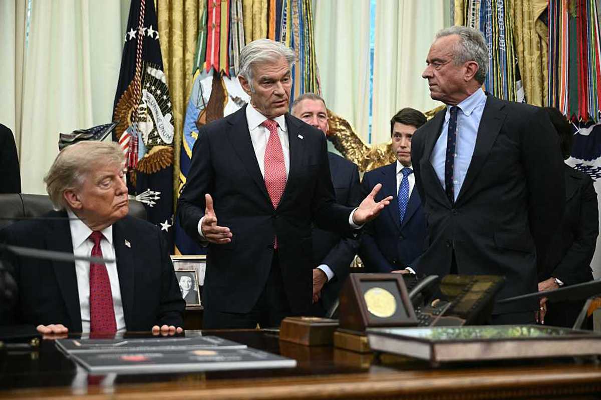 Dr Mehmet Oz speaking in the Oval Office, with Robert F. Kennedy Jr to the left of him and Donald Trump on his right, sitting at the Resolute Desk.