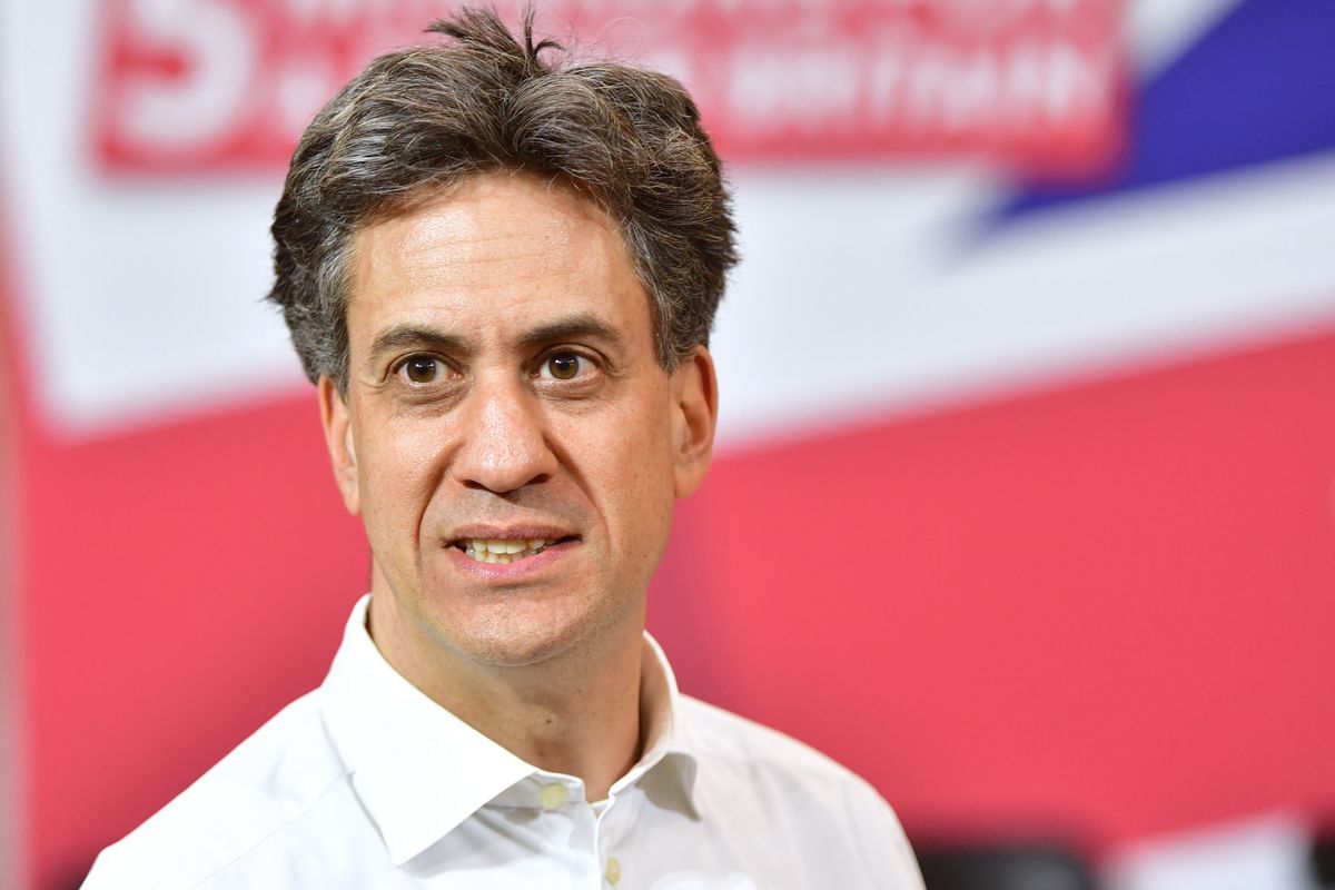 Ed Miliband, a white man with short black hair and a white collared shirt, with an out-of-focus Union Jack posterboard behind him.