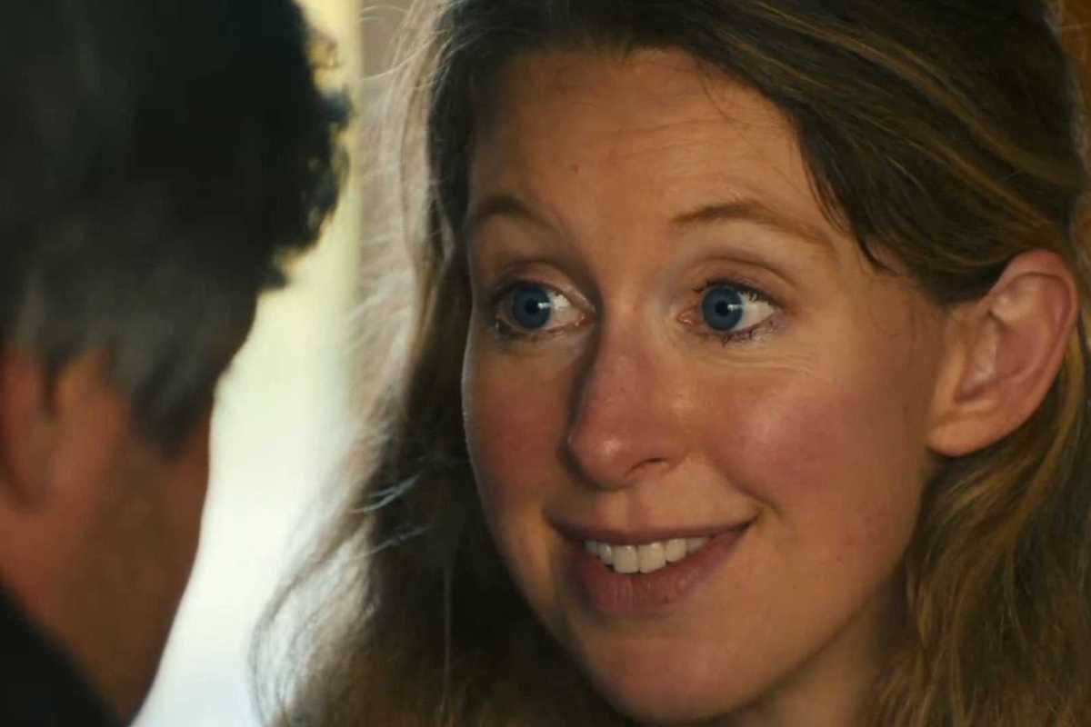 Elizabeth Holmes, a white woman with blue eyes and wavy blonde hair, smiles at a white man who has his back to the camera.