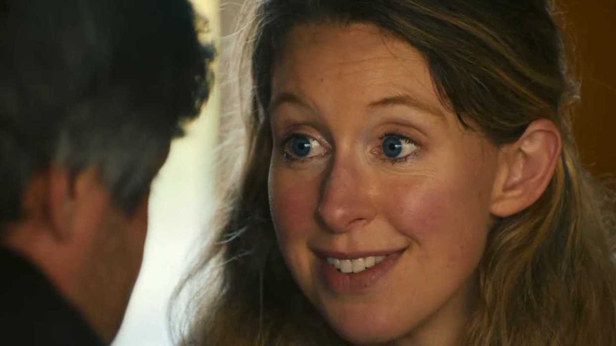 Elizabeth Holmes, a white woman with blue eyes and wavy blonde hair, smiles at a white man who has his back to the camera.