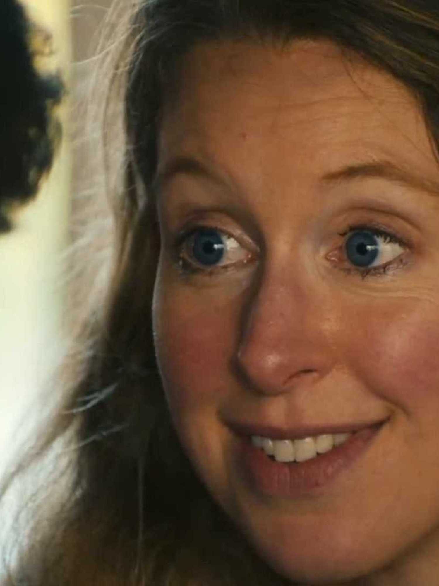 Elizabeth Holmes, a white woman with blue eyes and wavy blonde hair, smiles at a white man who has his back to the camera.