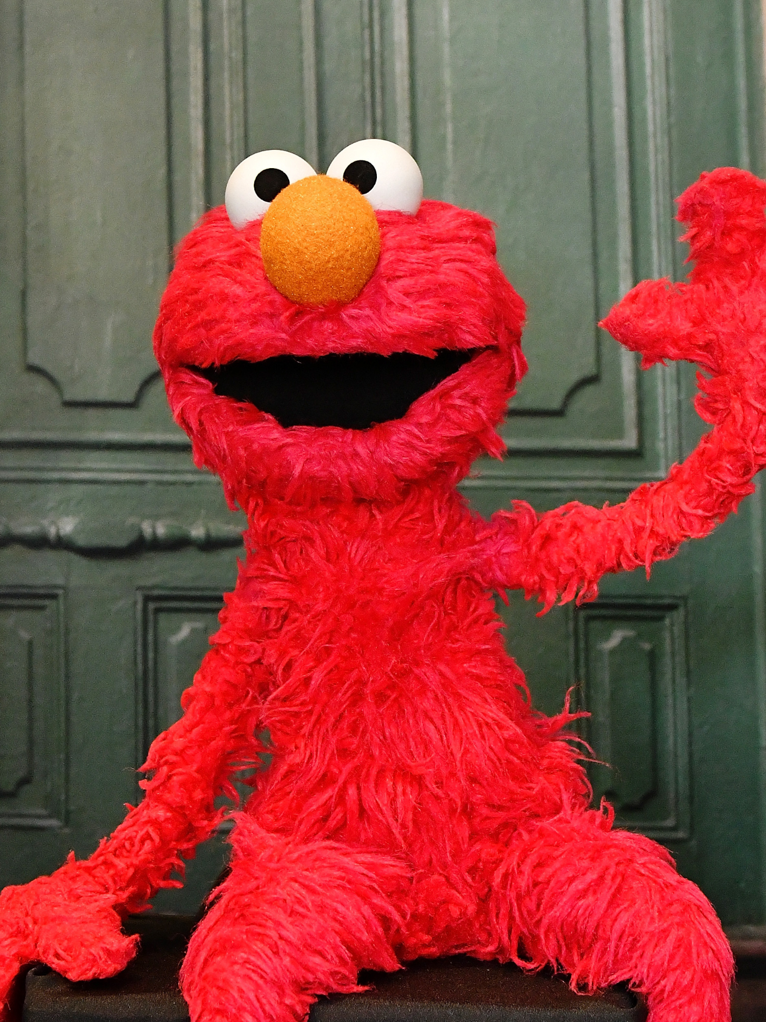 Elmo, the red Sesame Street puppet, smiling and waving outside a green door on Sesame Street.