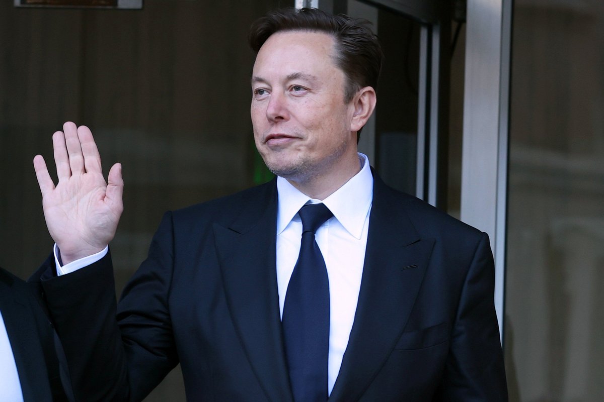 Elon Musk, a white man with short black hair and a black suit, holds up his right hand to wave at people off-camera.