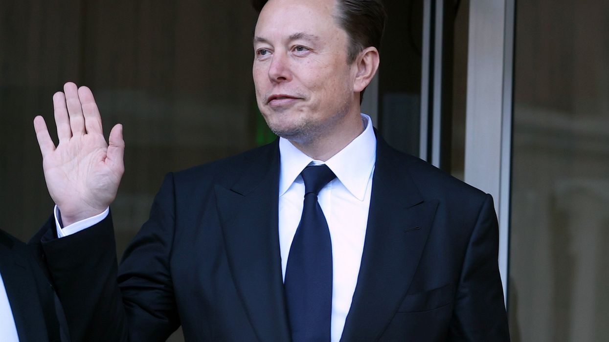 Elon Musk, a white man with short black hair and a black suit, holds up his right hand to wave at people off-camera.