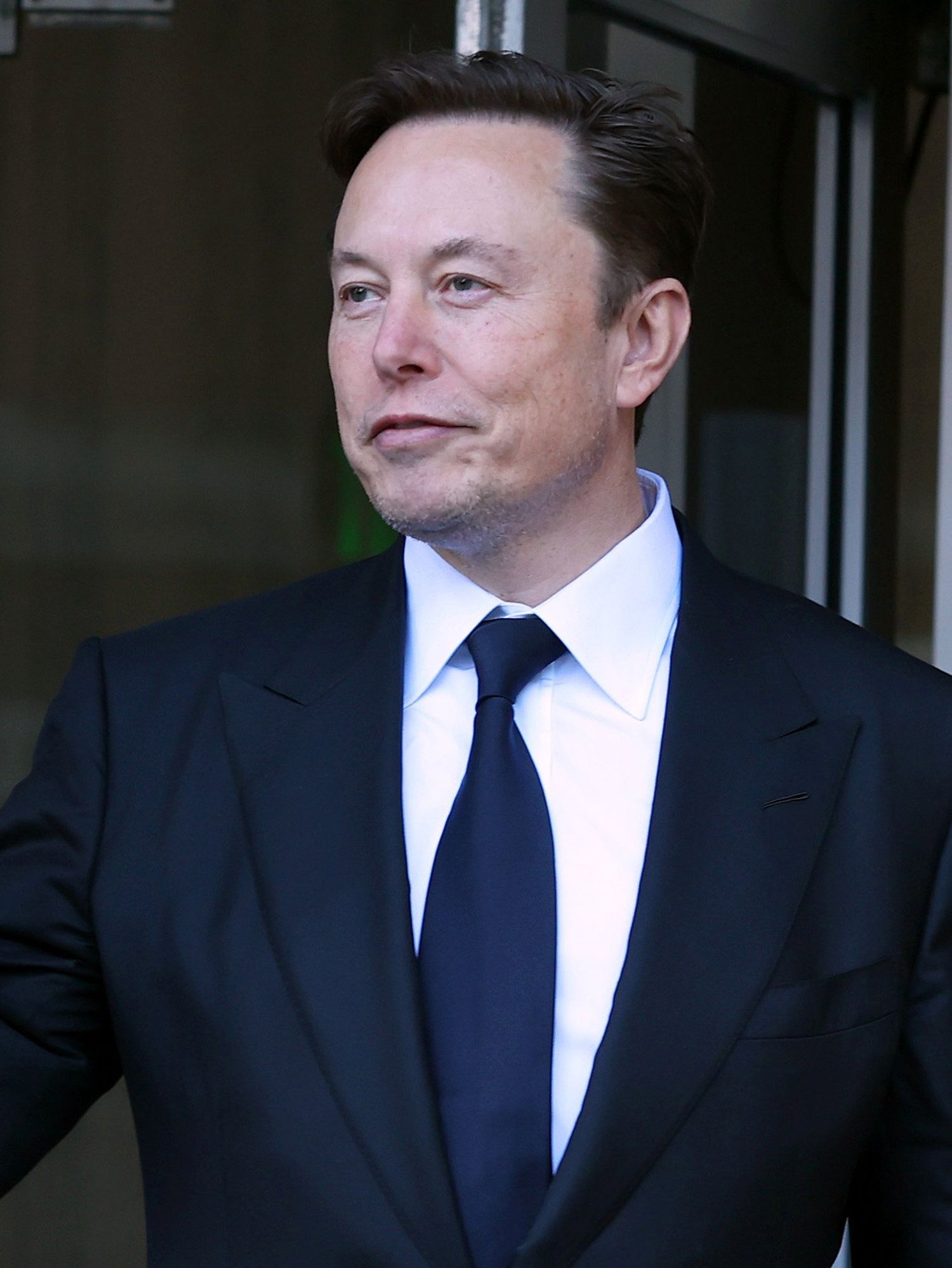 Elon Musk, a white man with short black hair and a black suit, holds up his right hand to wave at people off-camera.