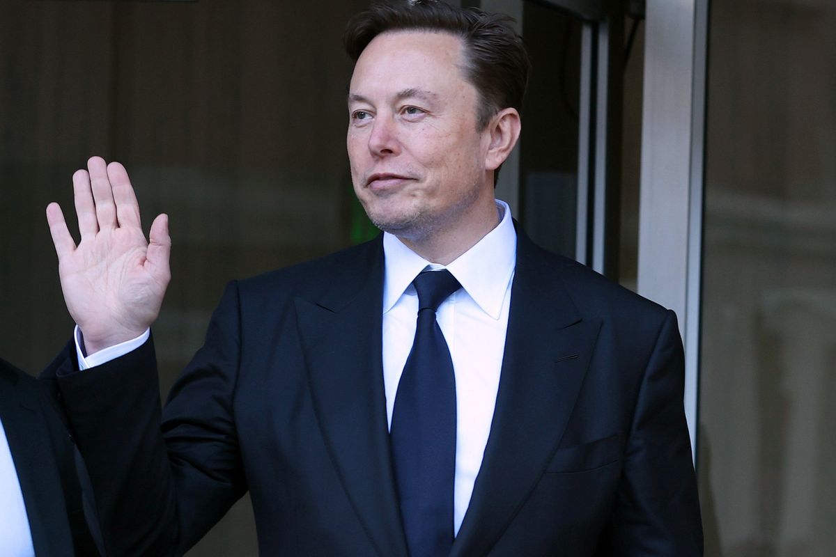 Elon Musk, a white man with short black hair and a black suit, waves at someone off-camera.