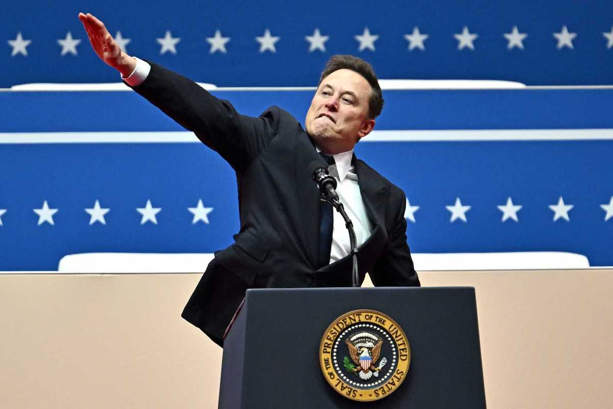 Elon Musk behind the US presidential lectern at Donald Trump's inauguration, gesturing to the crowd with a straight right arm pointing upwards.