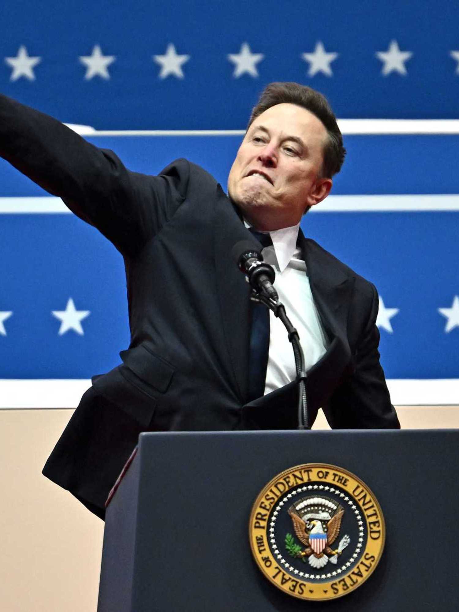 Elon Musk behind the US presidential lectern at Donald Trump's inauguration, gesturing to the crowd with a straight right arm pointing upwards.
