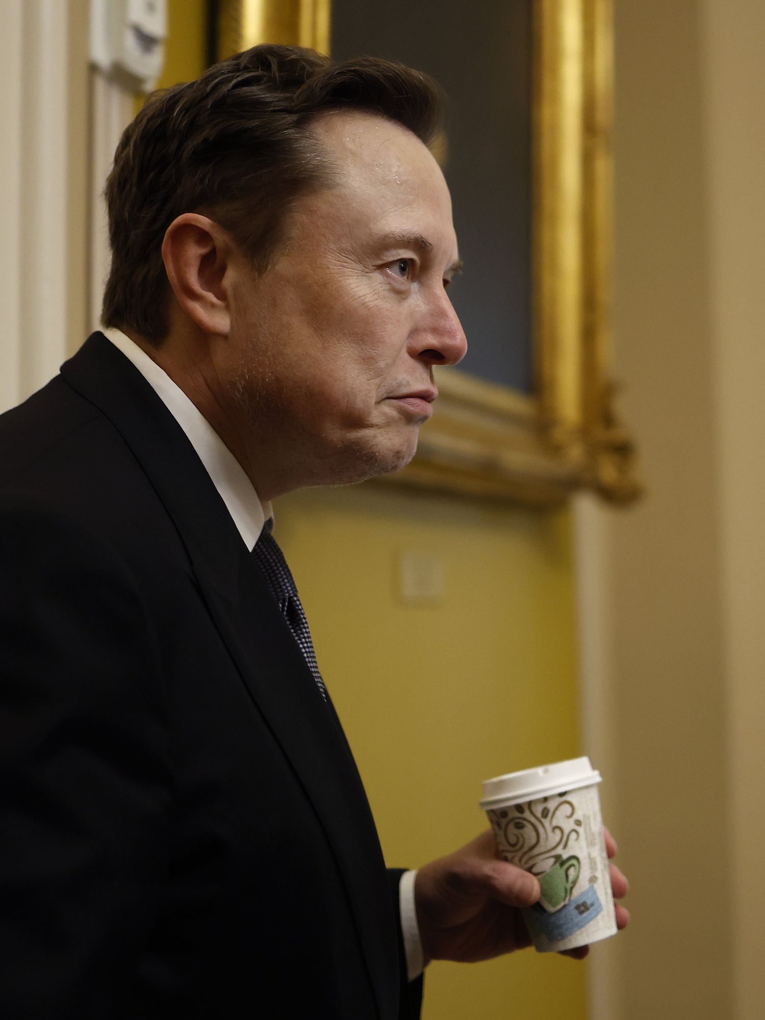 Elon Musk from the side on, holding a coffee cup and wearing a black suit.