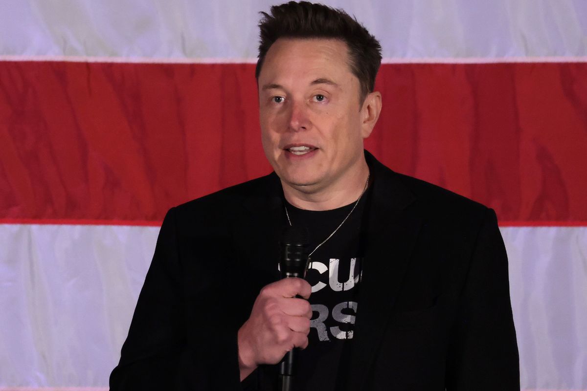 Elon Musk holds a microphone as he speaks in front of an American flag.
