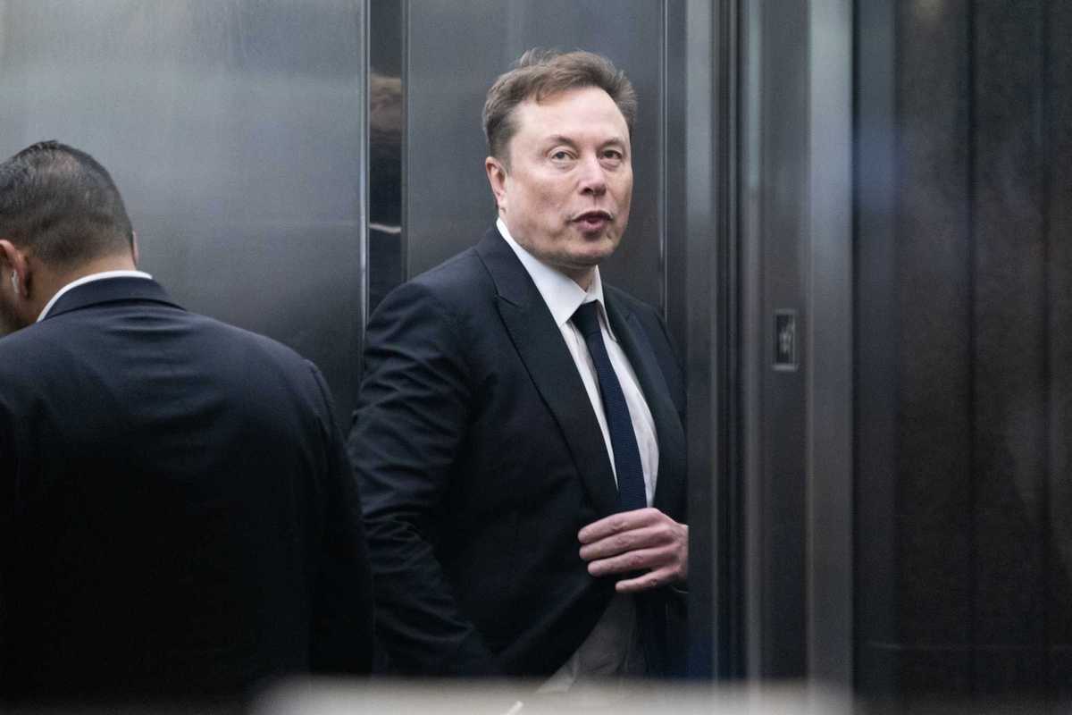 Elon Musk in a black suit and tie against a metal wall.