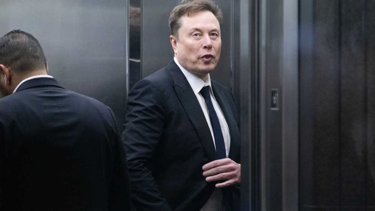Elon Musk in a black suit and tie against a metal wall.