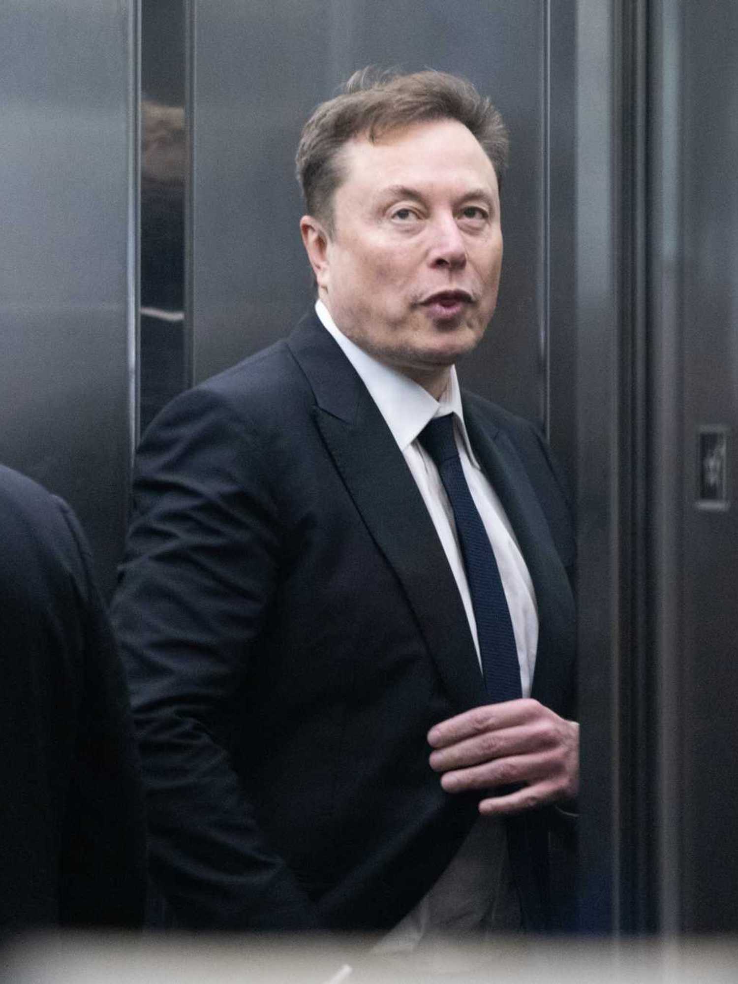Elon Musk in a black suit and tie against a metal wall.