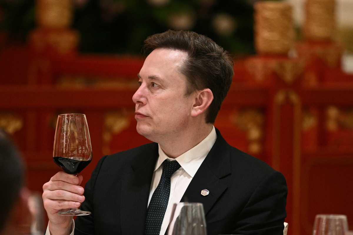 Elon Musk in a black suit and tie, holding a glass of red wine in his right hand.