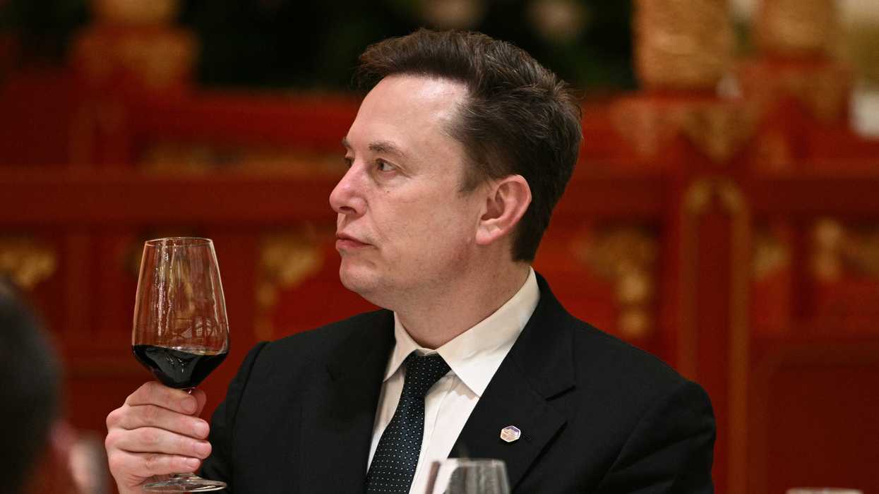 Elon Musk in a black suit and tie, holding a glass of red wine in his right hand.