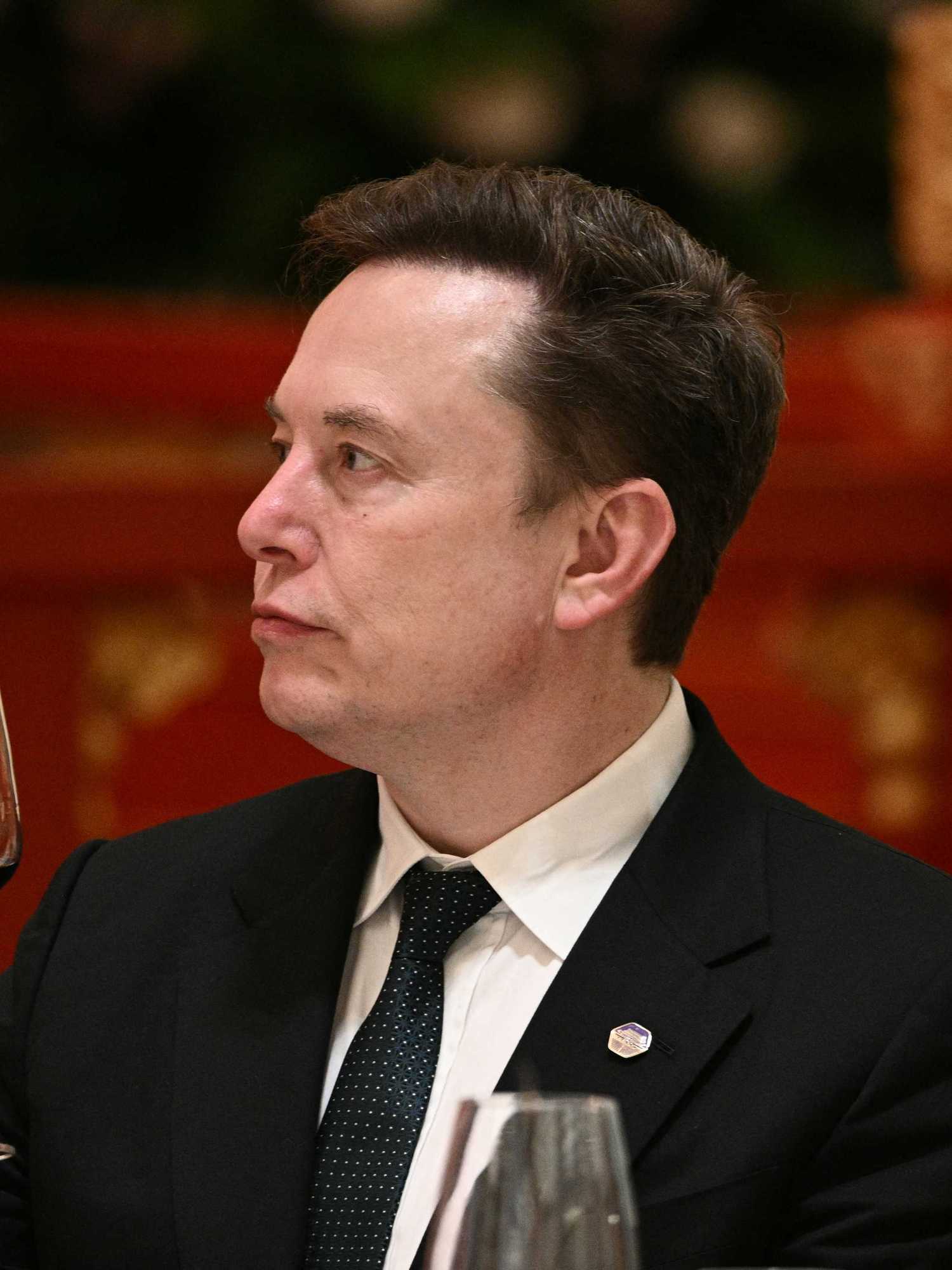 Elon Musk in a black suit and tie, holding a glass of red wine in his right hand.