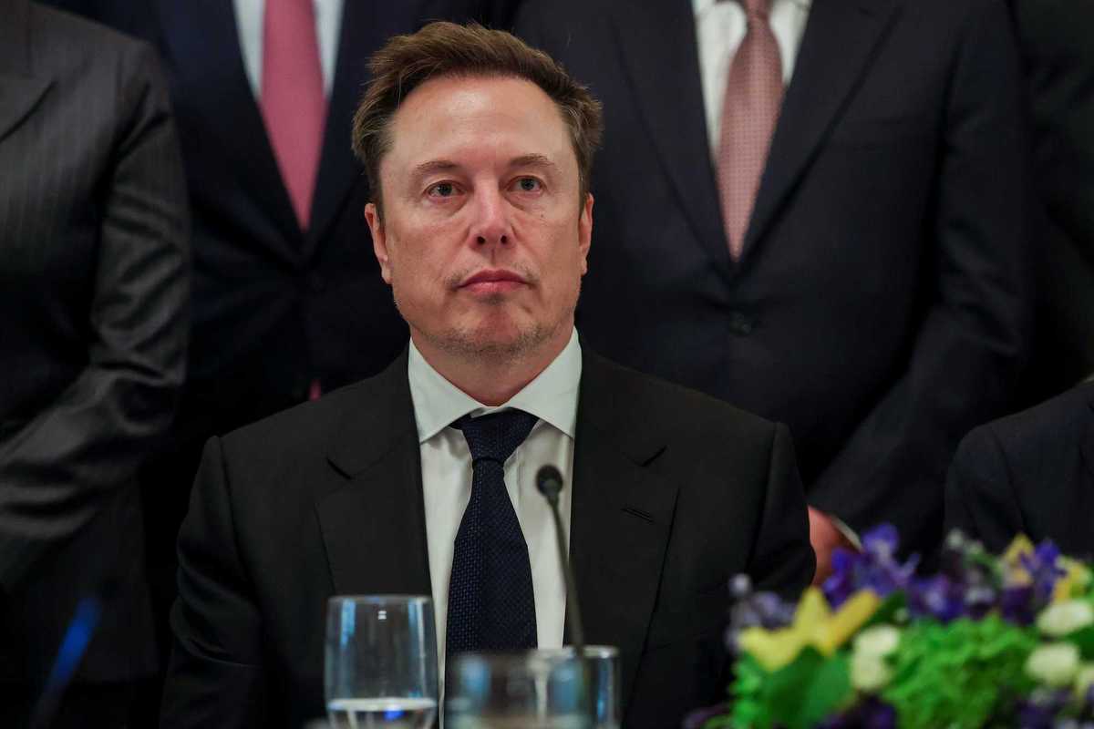 Elon Musk, in a black suit and tie, sitting at a table, with a neutral expression on his face.