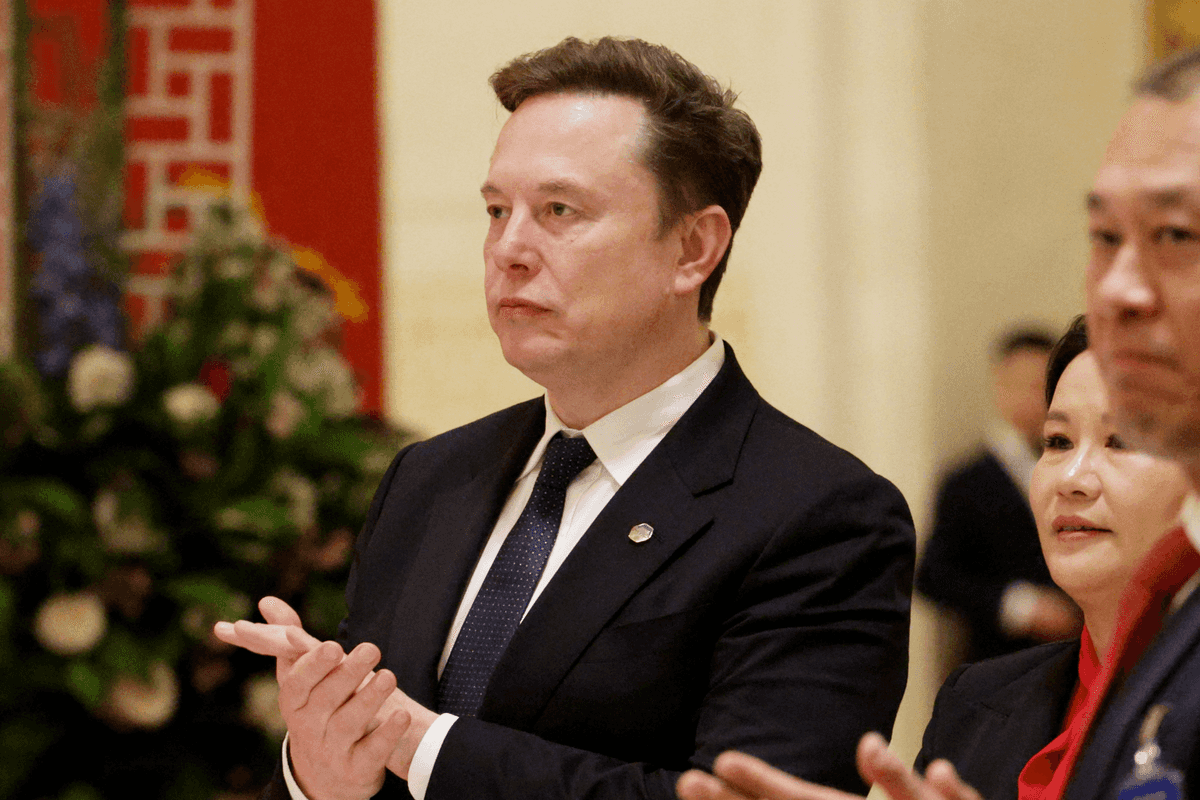Elon Musk in a black suit clapping his hands with a neutral expression on his face.