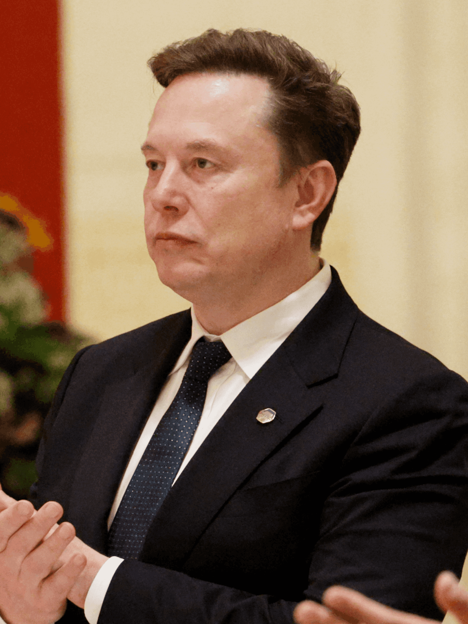Elon Musk in a black suit clapping his hands with a neutral expression on his face.