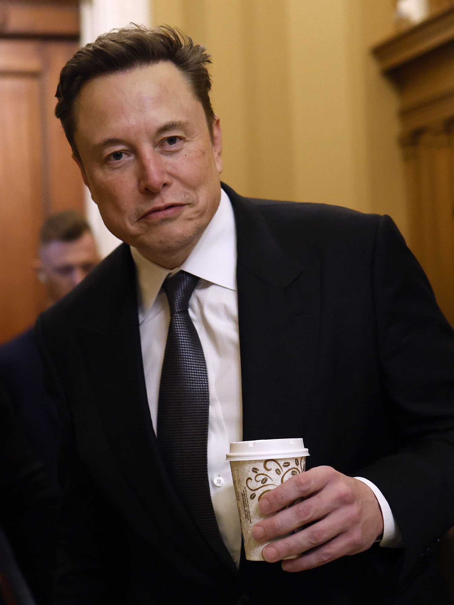 Elon Musk in a black suit holding a coffee cup.
