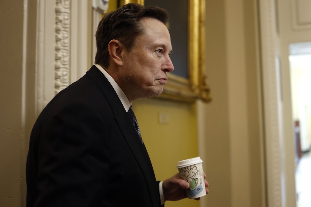 Elon Musk in a black suit holding a coffee cup.