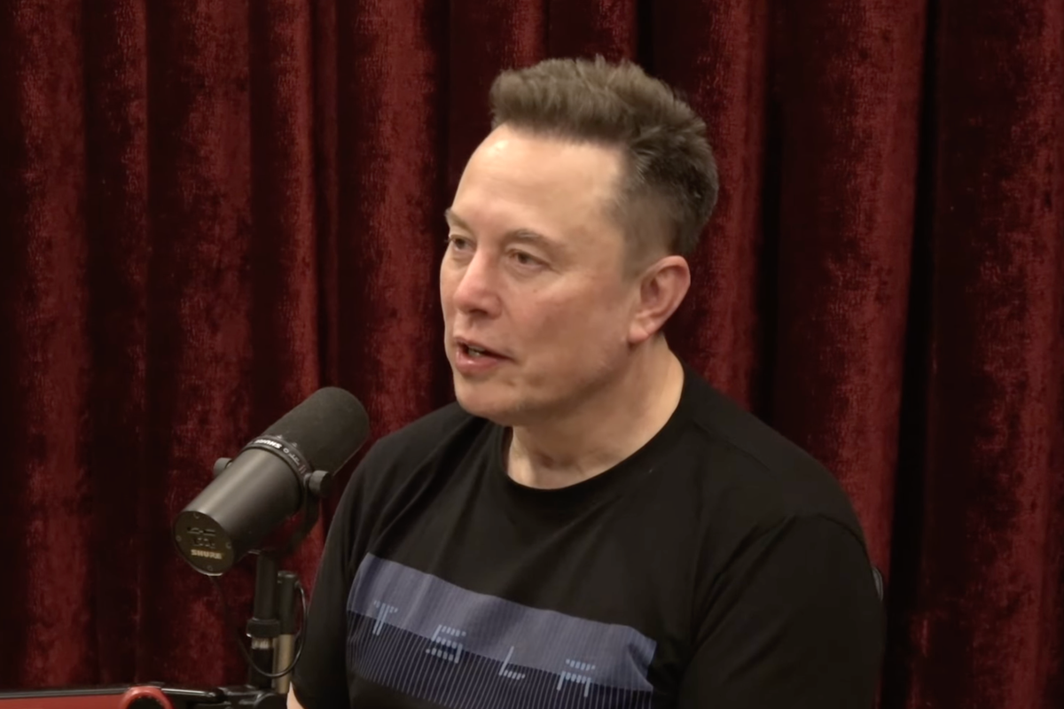Elon Musk in a podcast studio, speaking into a microphone, with a red curtain behind him.