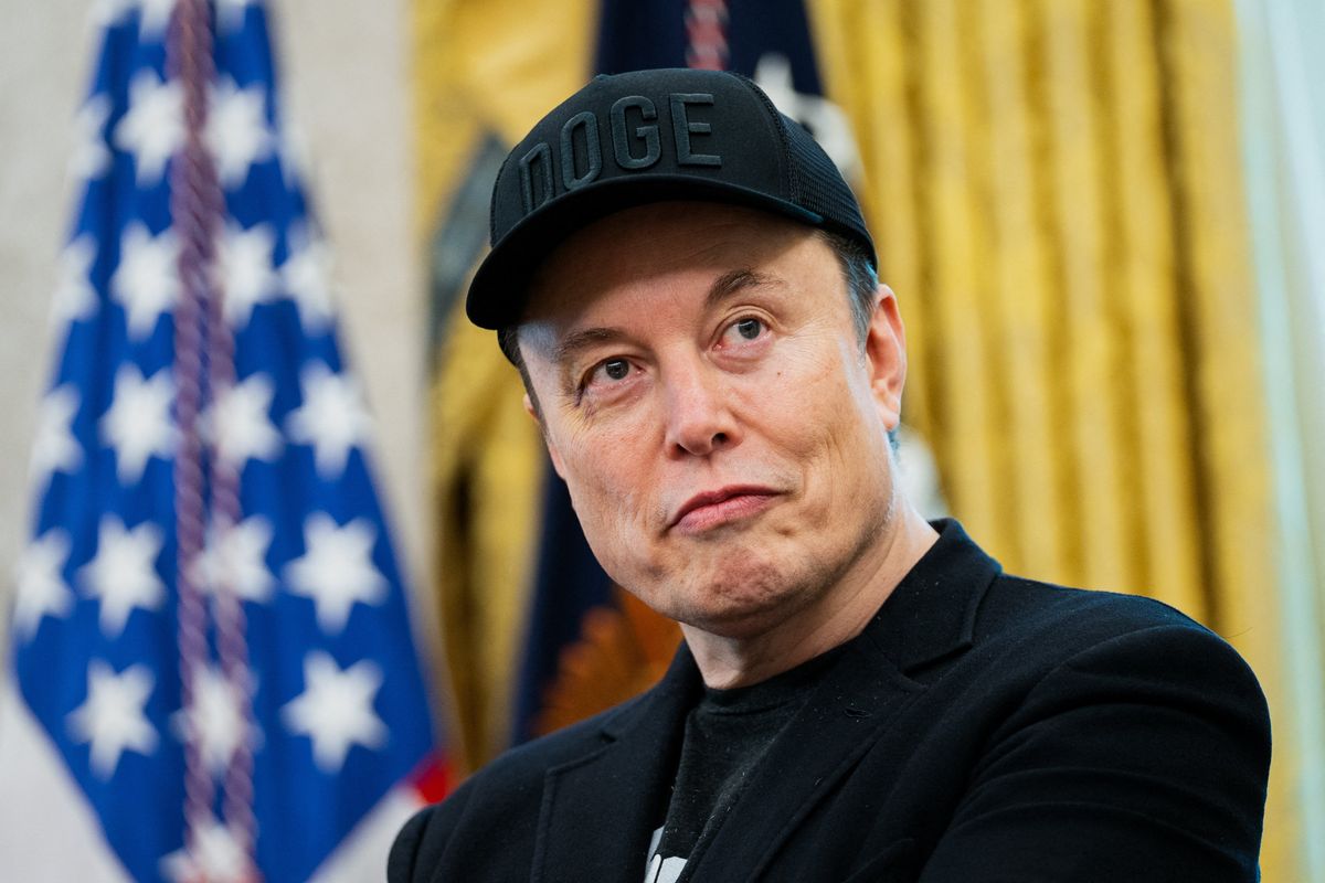 Elon Musk looks to his left, wearing a black DOGE cap and a black suit jacket. He has a neutral facial expression and is in the Oval Office.
