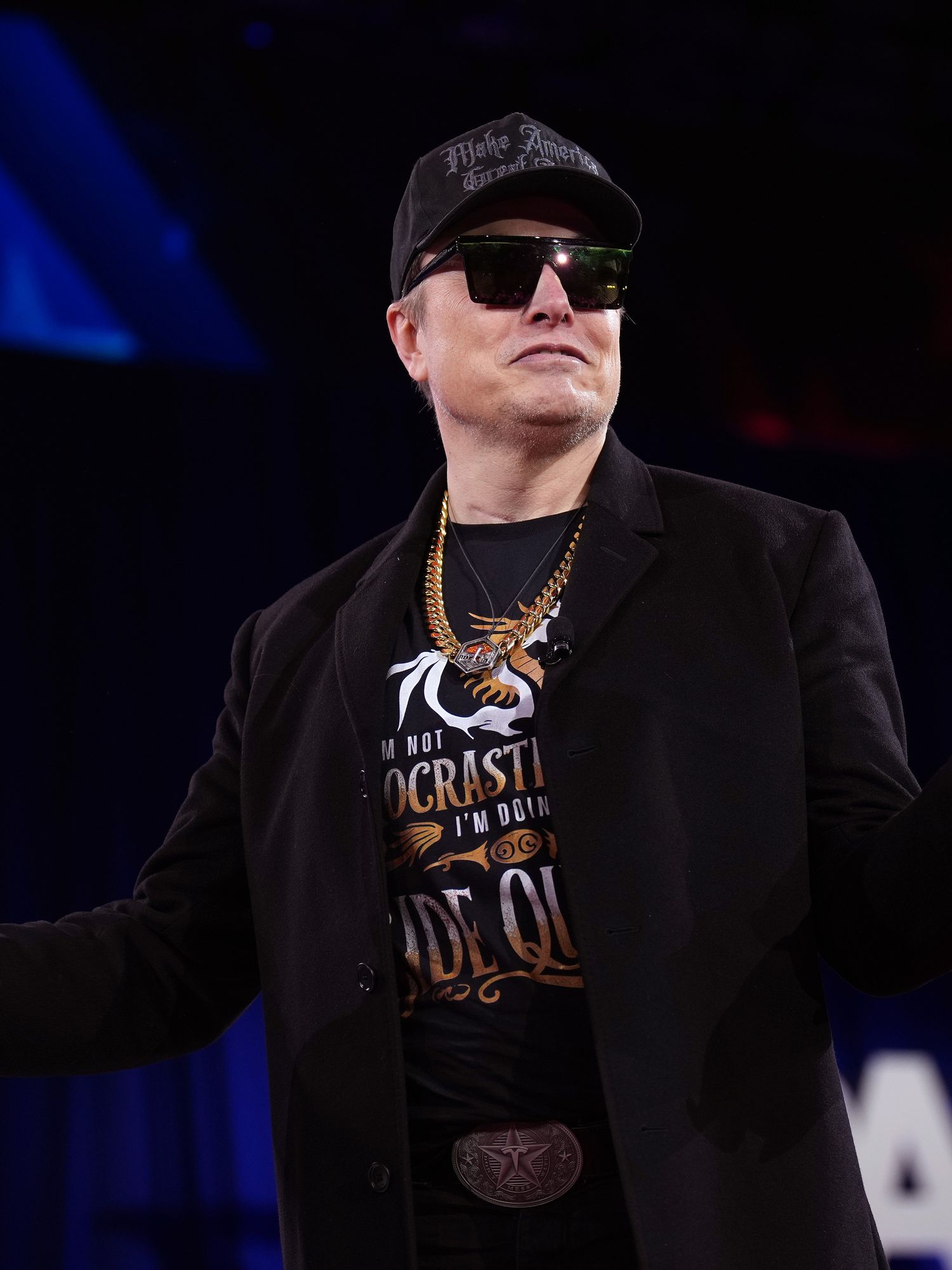 Elon Musk on stage at CPAC, wearing black sunglasses and all-black clothing, smiling at the crowd and shrugging with upturned palms.