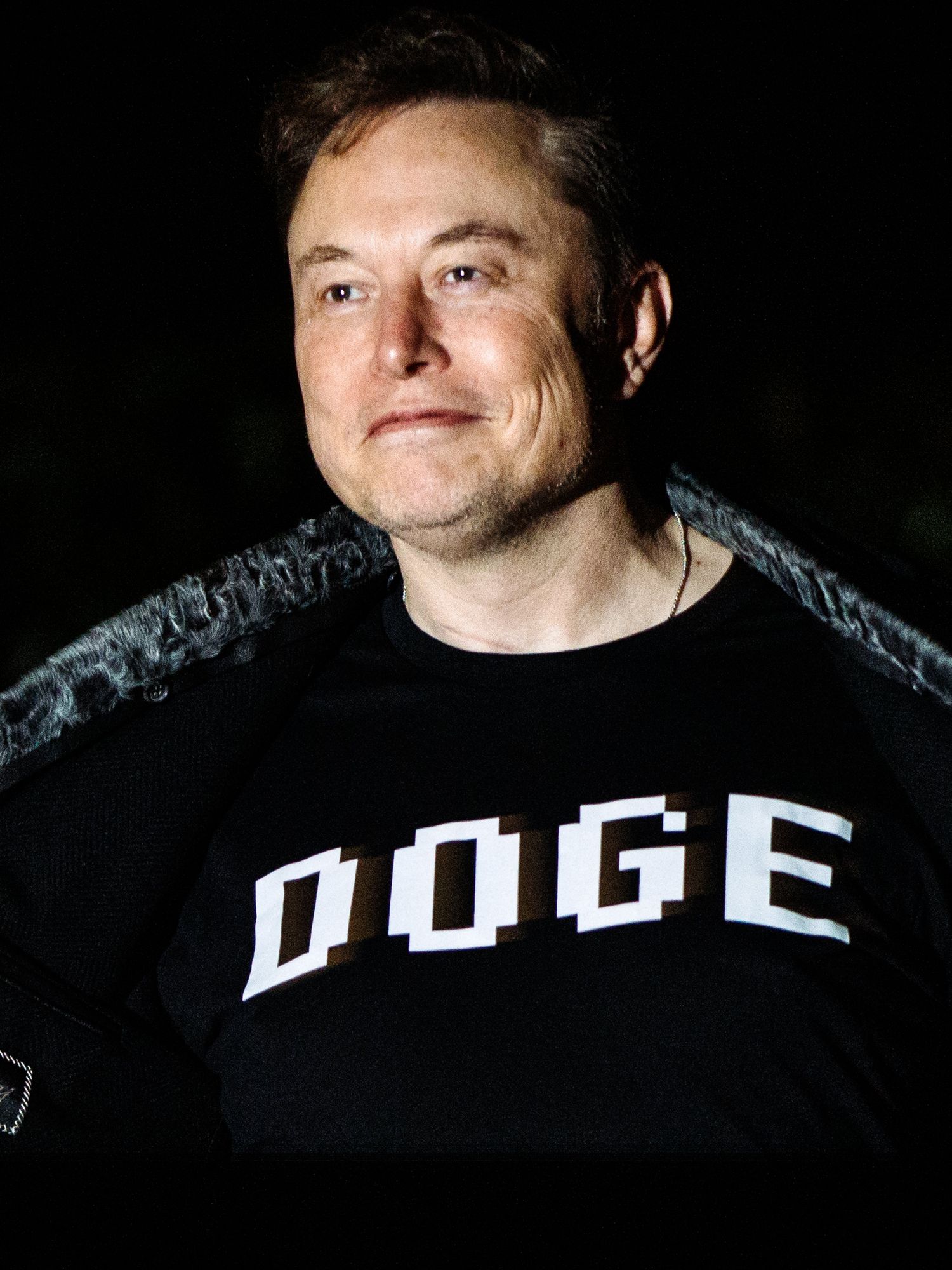 Elon Musk opens up his black jacket to reveal a black T-shirt with the word 'Doge' on it in white text. He is smirking.