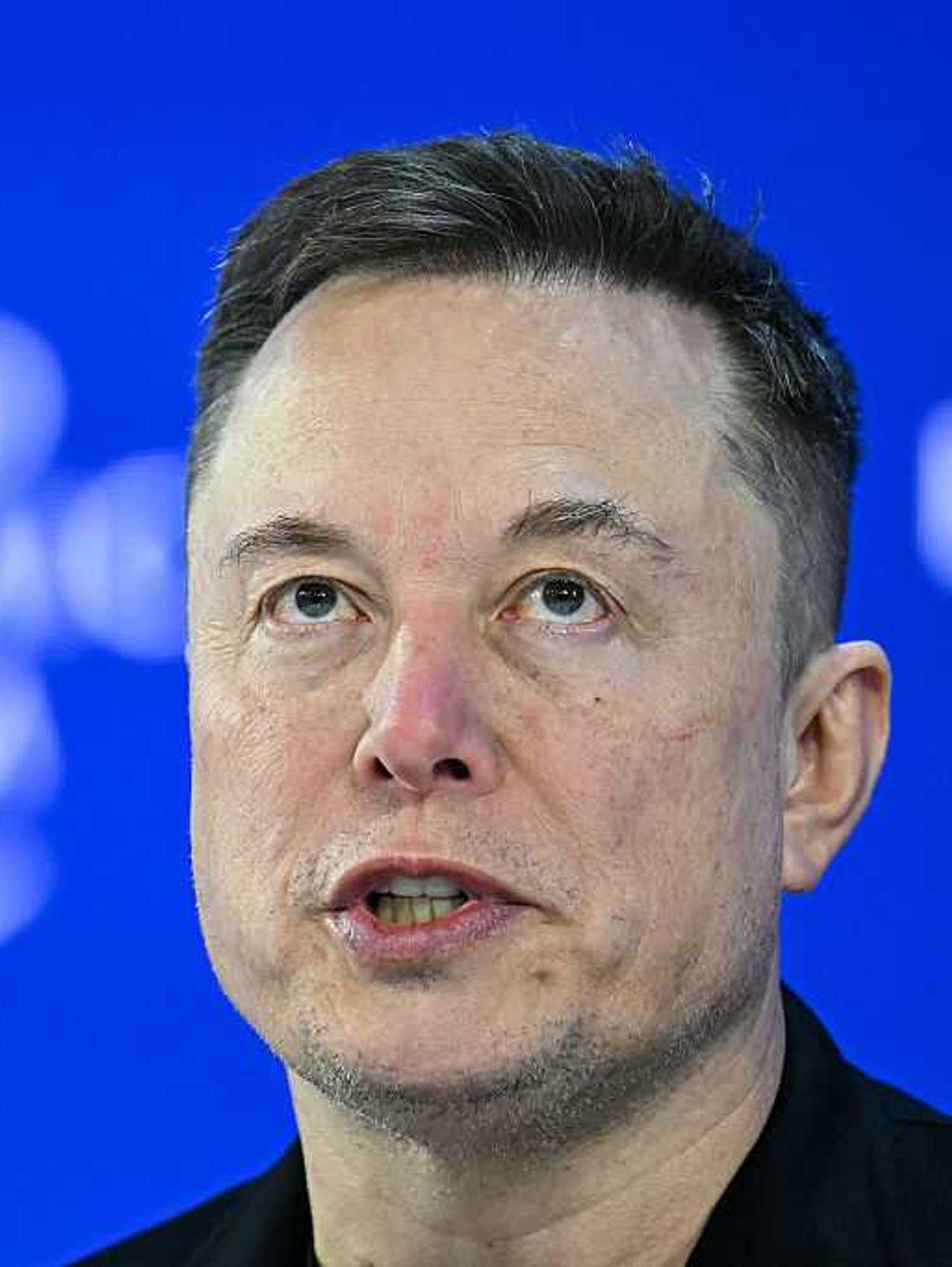 Elon Musk talking at the World Economic Forum in front of a blue posterboard background, with the white 'World Economic Forum' text out of focus.