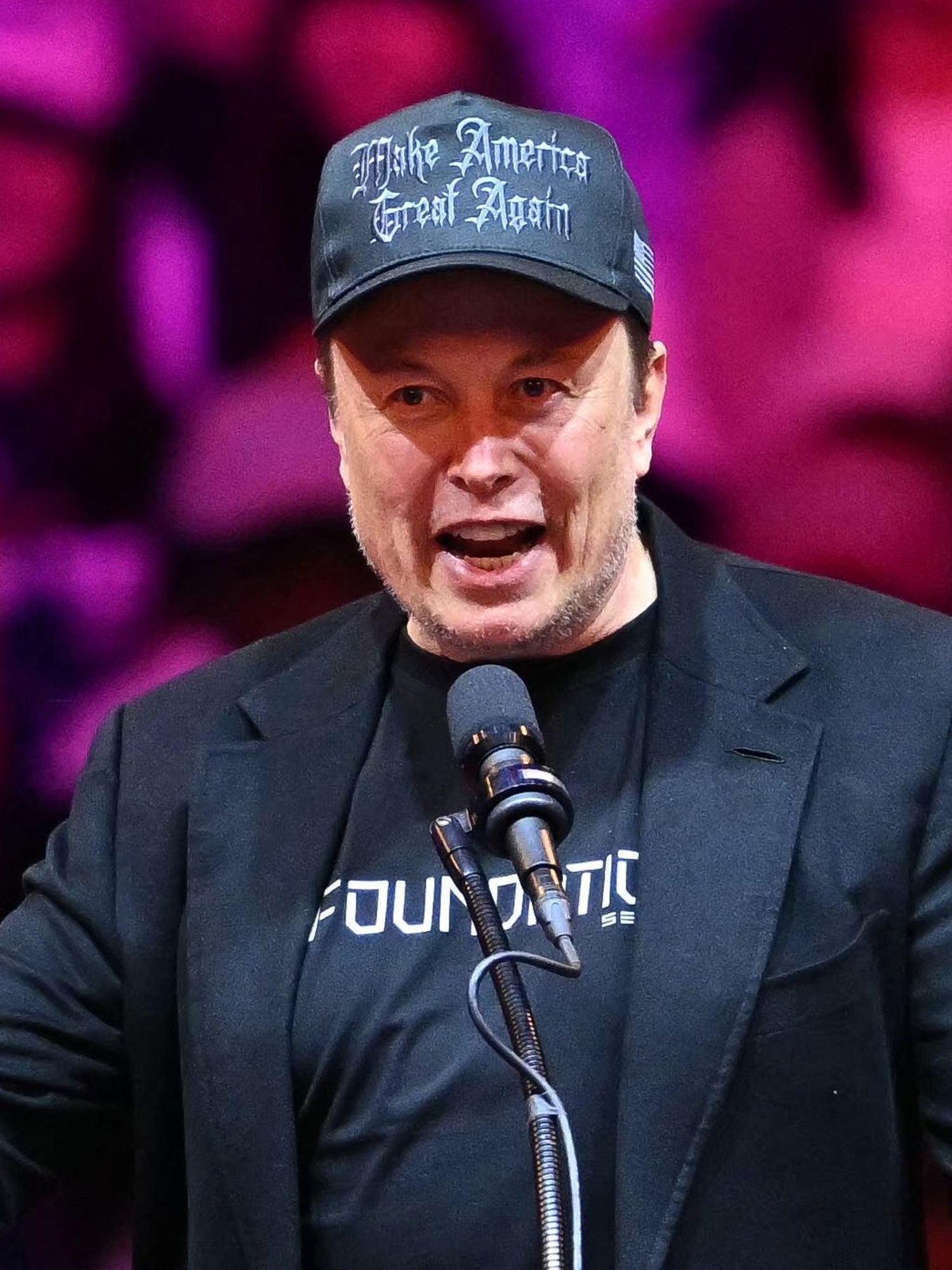 Elon Musk wearing a black Make America Great Again hat and speaking at a Donald Trump rally.