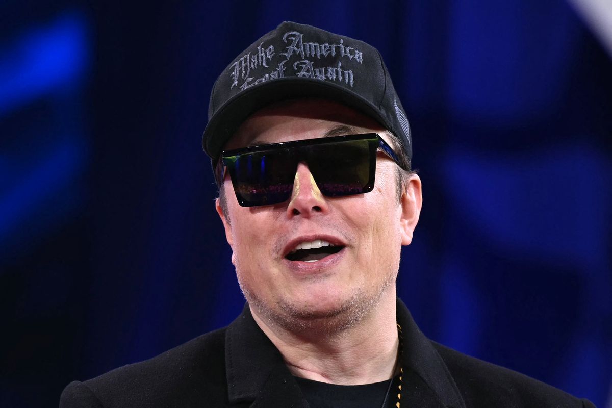 Elon Musk wearing a black suit, black sunglasses and a black 'Make America Great Again' cap.