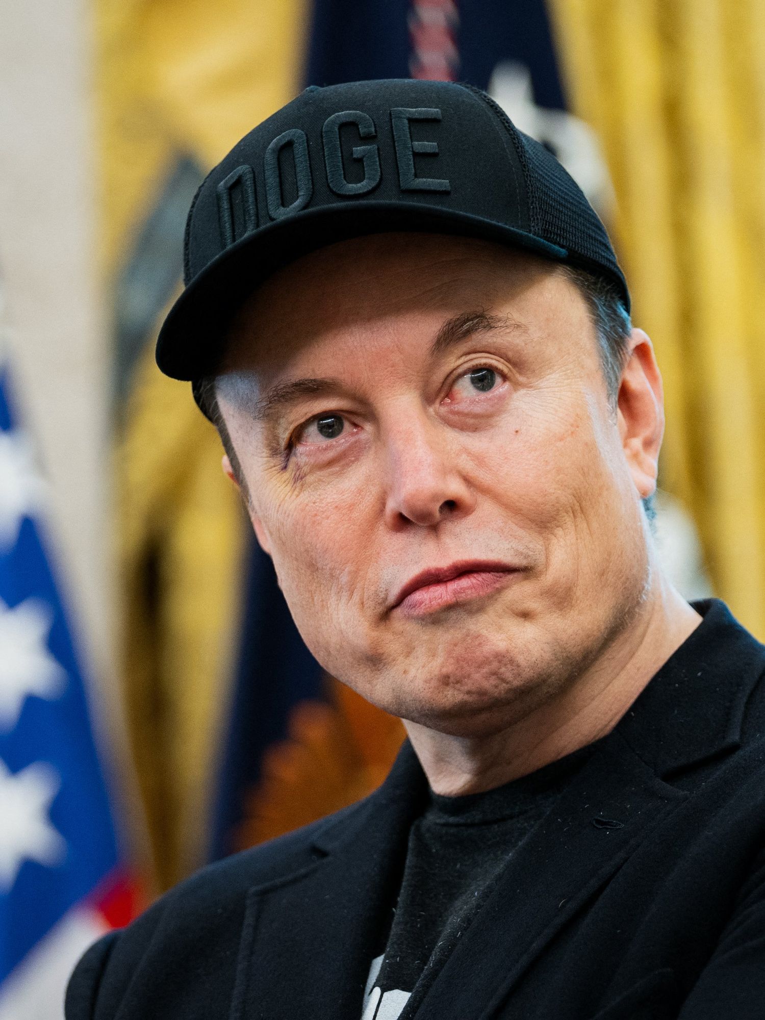 Elon Musk with a neutral expression on his face, wearing a black Doge cap and black suit jacket. He's in the Oval Office.
