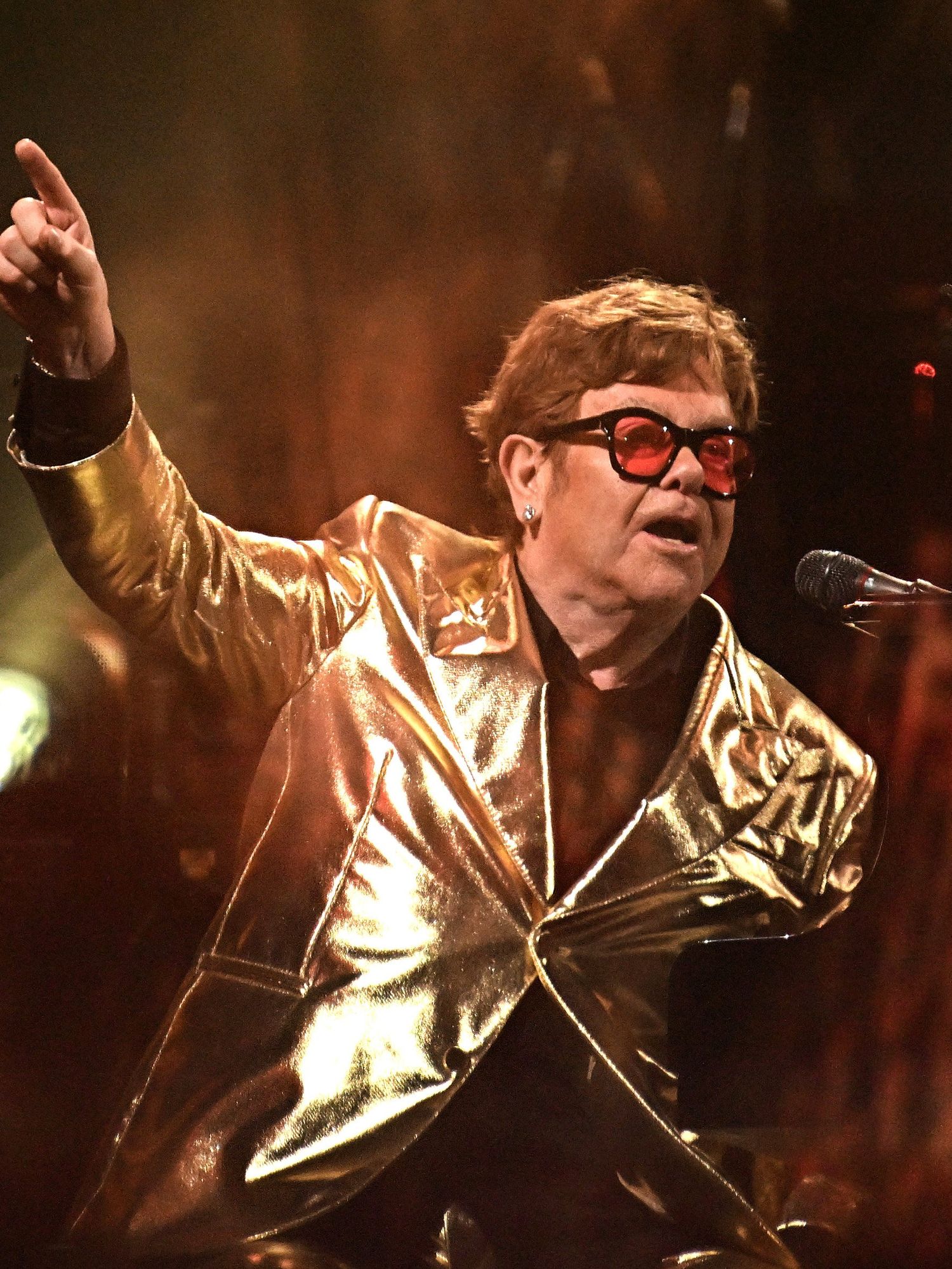 Elton John, a white man with short brown hair, red sunglasses and a gold suit, points his right index finger upwards as he sings on stage at Glastonbury.
