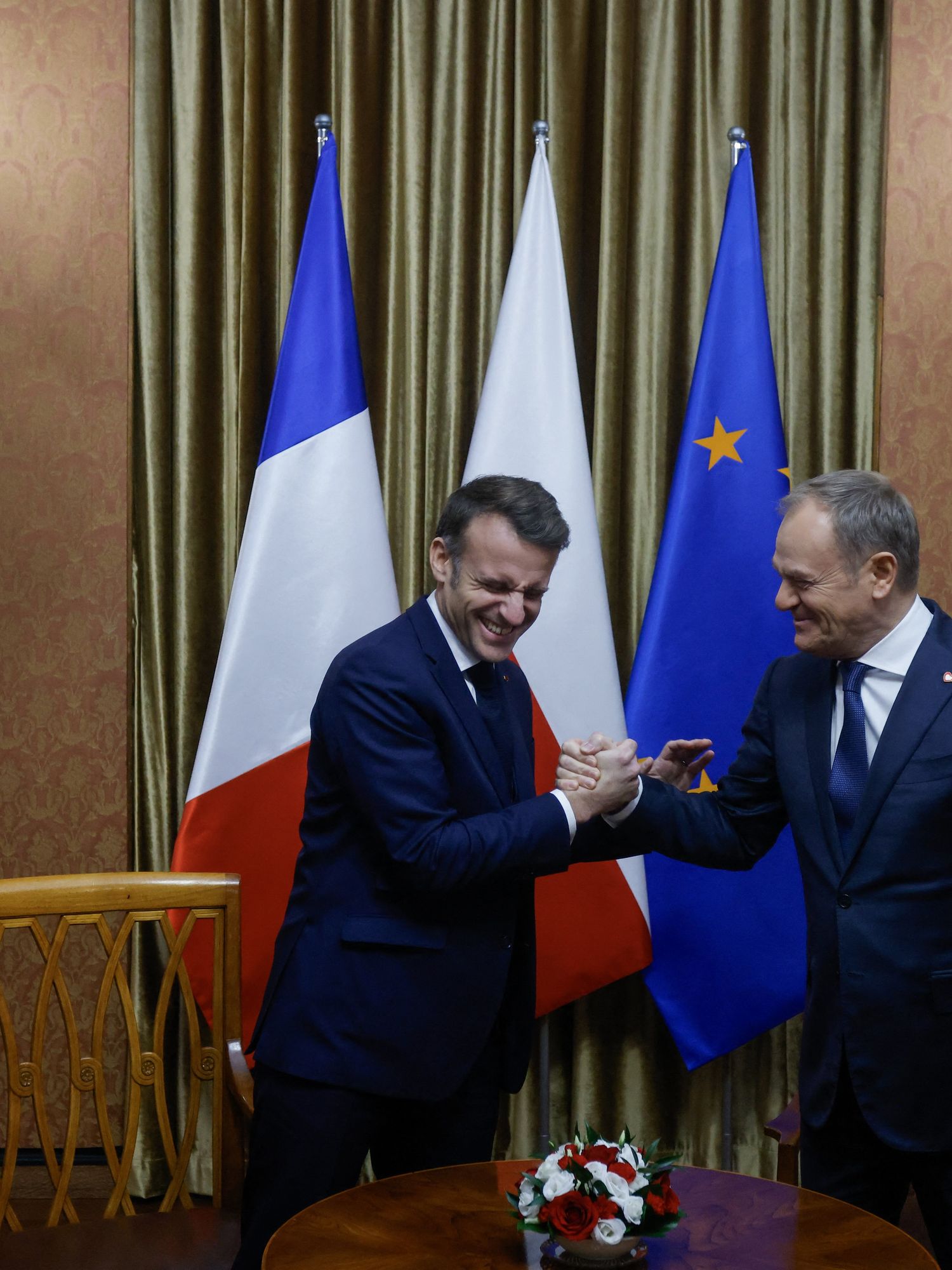 Emmanuel Macron and Donald Tusk laugh as they share a handshake in a meeting room.