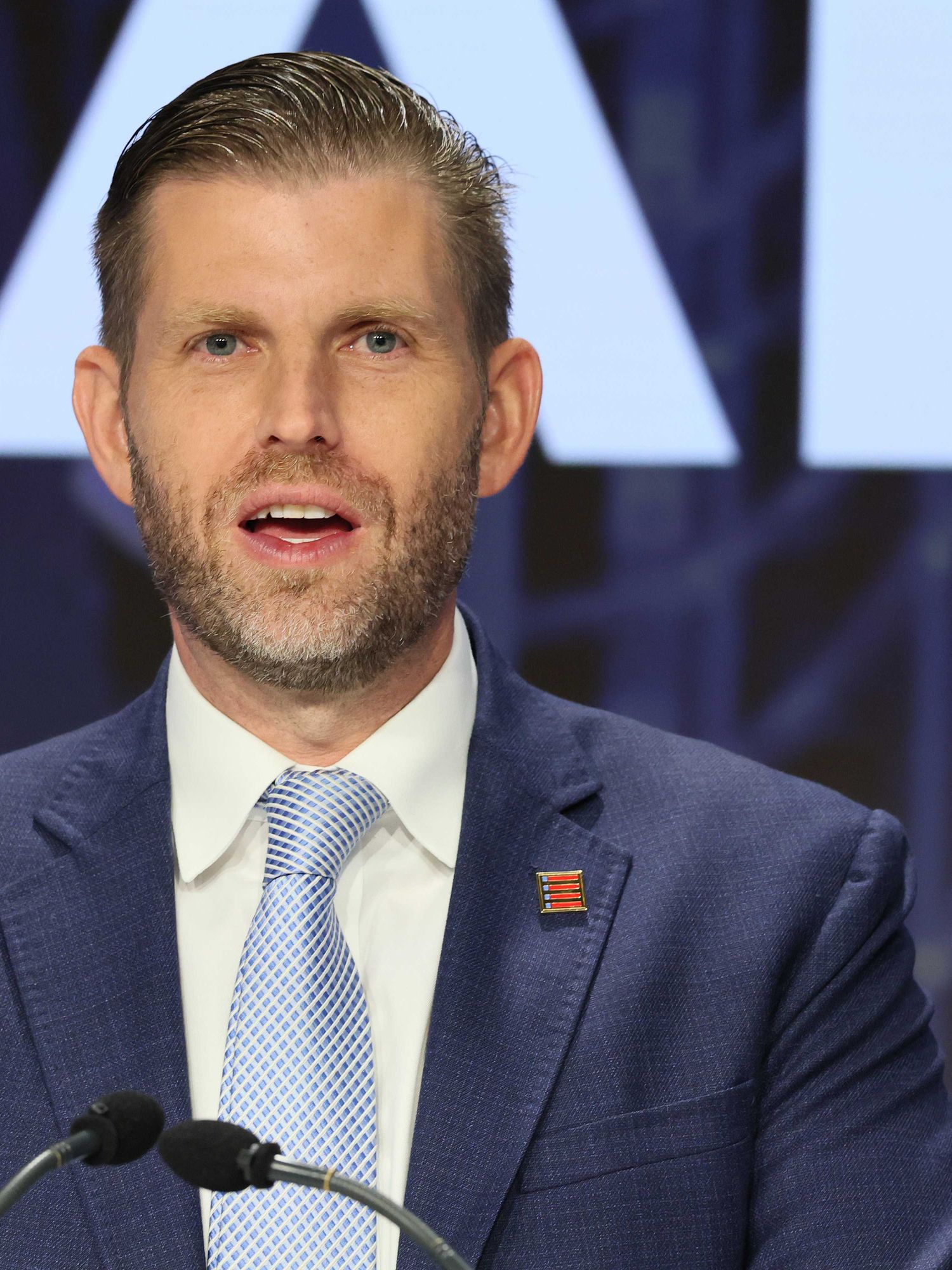 Eric Trump speaks behind a lectern, wearing a blue suit.