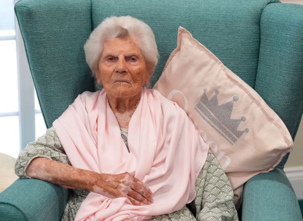 Ethel Caterham seated in an armchair, on a cushion which reads 'Queen'
