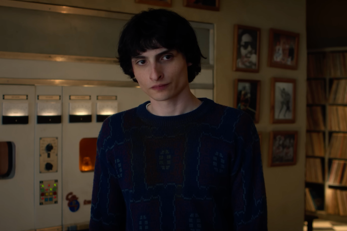 Finn Wolfhard as Mike Wheeler in Stranger Things. He has short black hair and a blue jumper, and is standing in a room with radio machinery and framed pictures behind him.