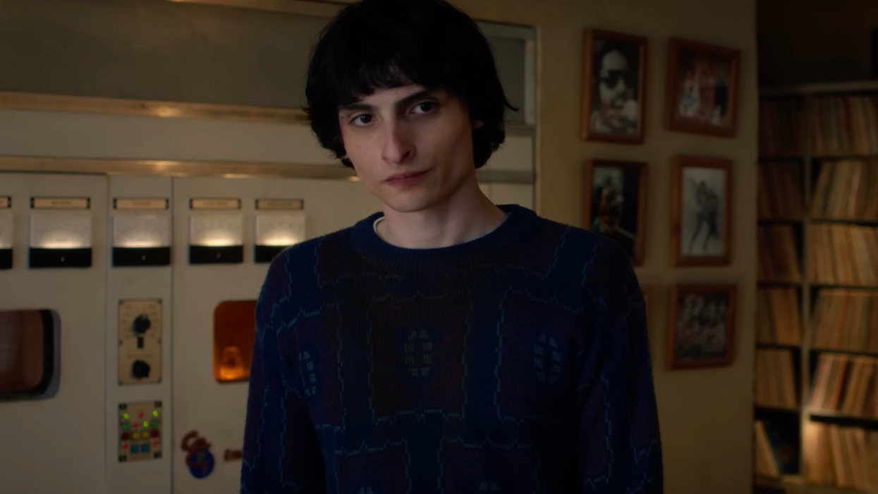 Finn Wolfhard as Mike Wheeler in Stranger Things. He has short black hair and a blue jumper, and is standing in a room with radio machinery and framed pictures behind him.