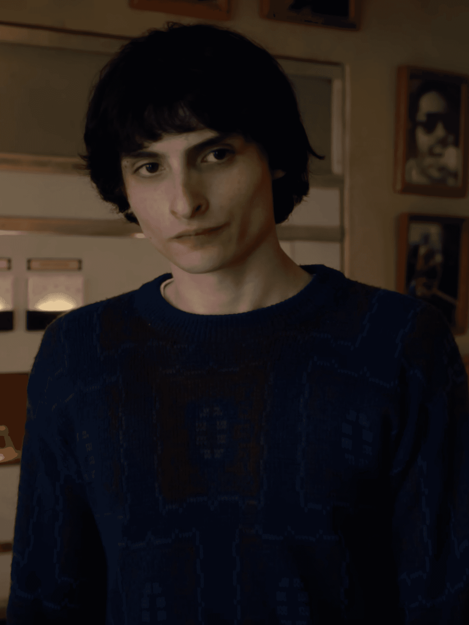 Finn Wolfhard as Mike Wheeler in Stranger Things. He has short black hair and a blue jumper, and is standing in a room with radio machinery and framed pictures behind him.