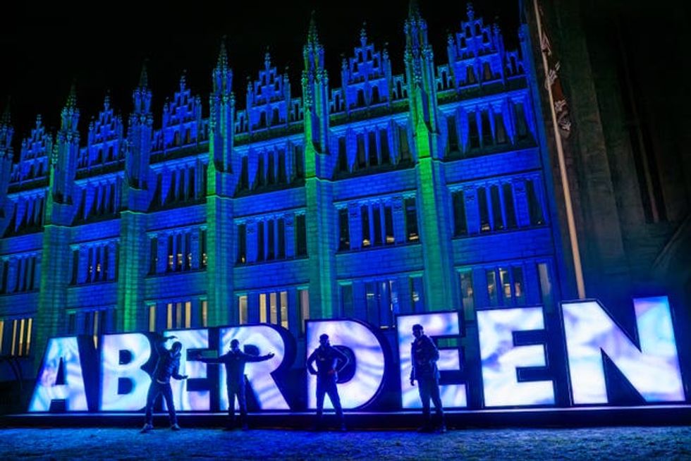 Four people standing in front of giant letters which spell Aberdeen, with a building lit up in blue behind it