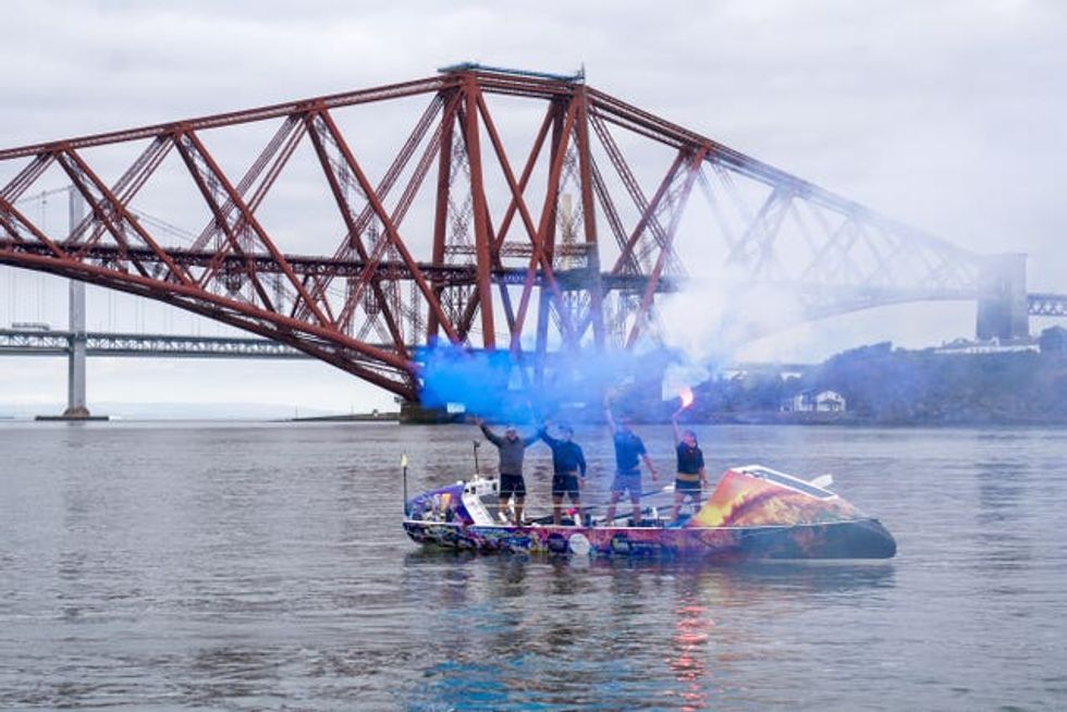 Four rowers in a boat stand while waving flares, in front of the Forth Bridge