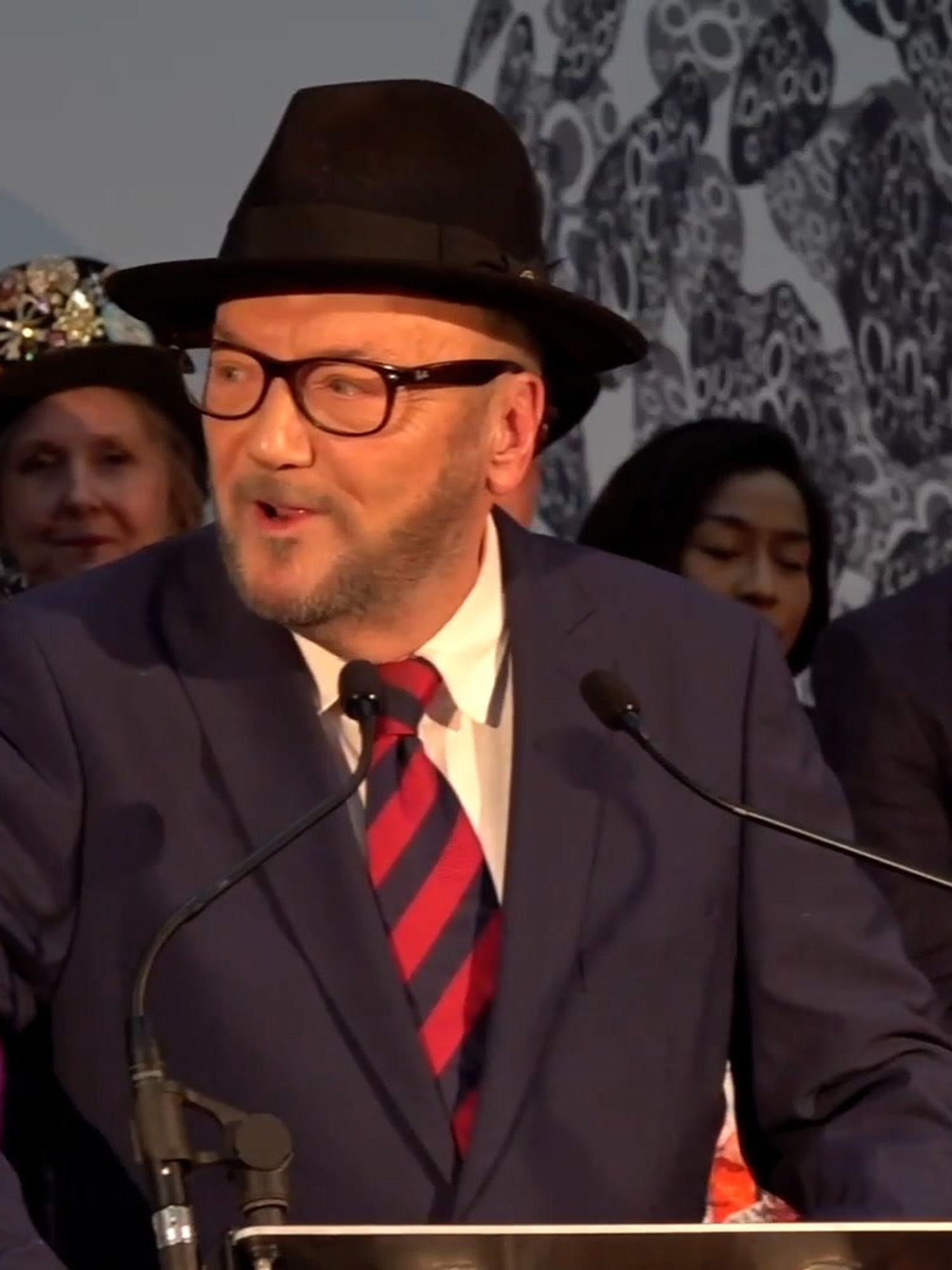 George Galloway, a bearded white man with glasses, a grey beard, black suit and fedora, speaks in front of a lectern.