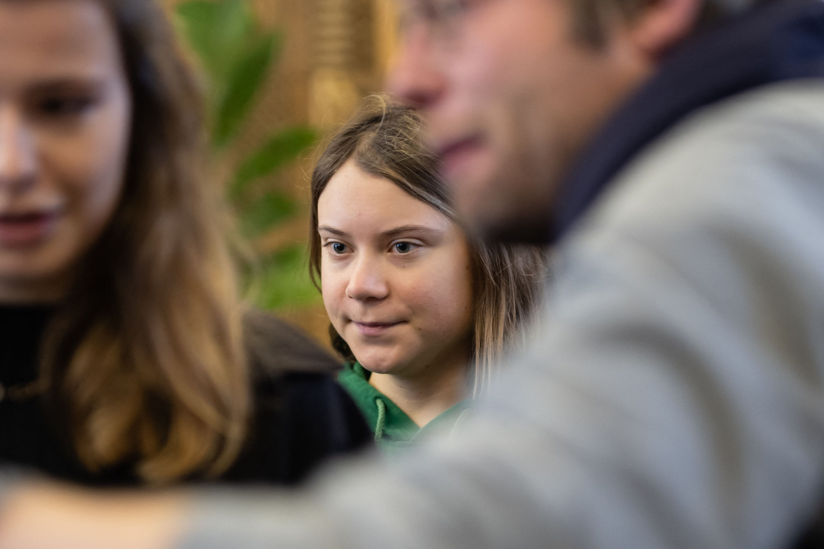Greta Thunberg, a young white woman with short blonde hair and a green jumper has a neutral expression on her face. Blurred in the foreground of the photo are a man and woman talking.