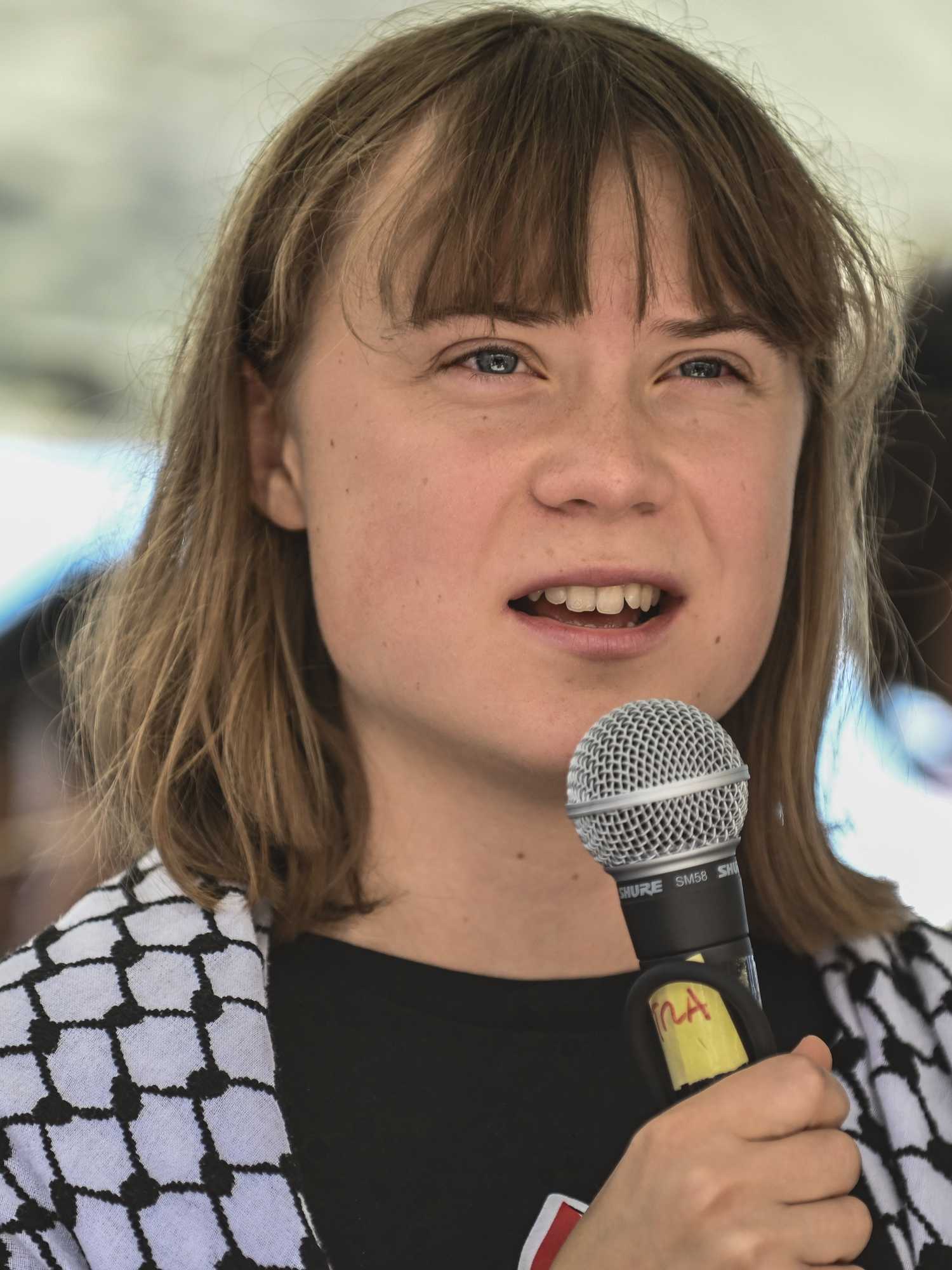 Greta Thunberg speaks into a microphone, with a keffiyeh wrapped around her shoulders.