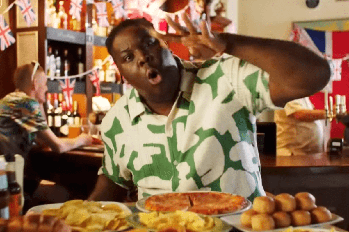 Hammed Animashaun, a Black man with short black hair and a green and white shirt, holds up four fingers while standing behind four plates of food in a pub.
