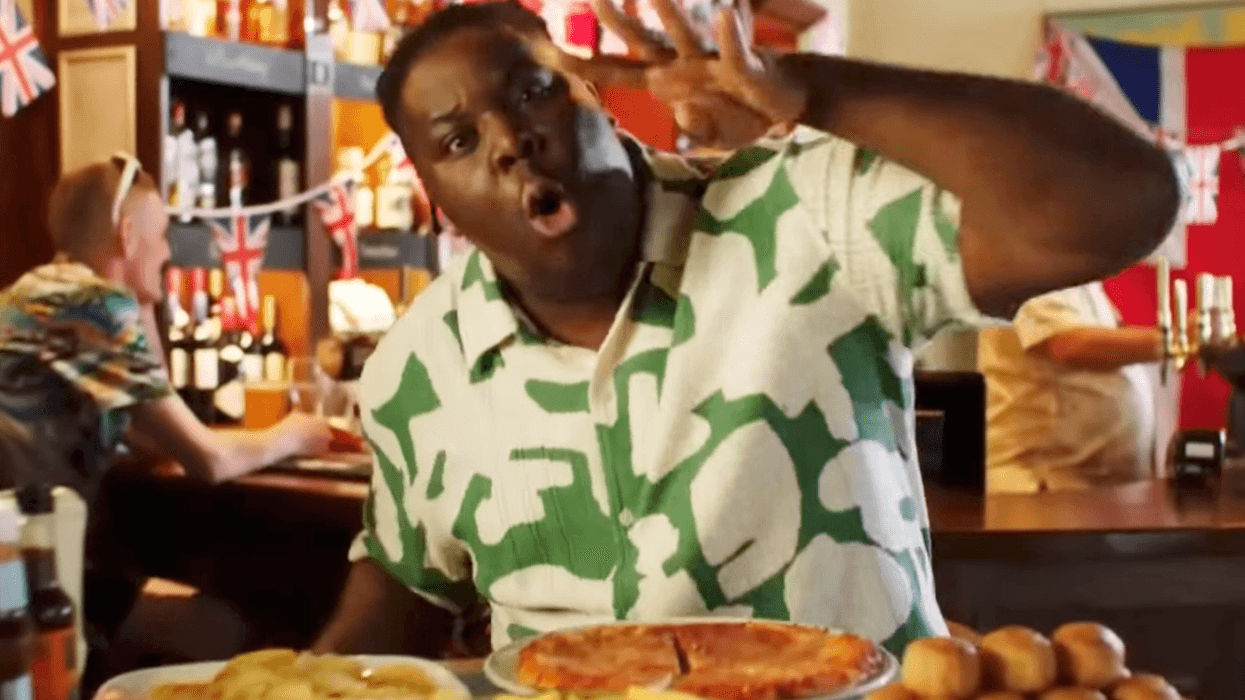 Hammed Animashaun, a Black man with short black hair and a green and white shirt, holds up four fingers while standing behind four plates of food in a pub.