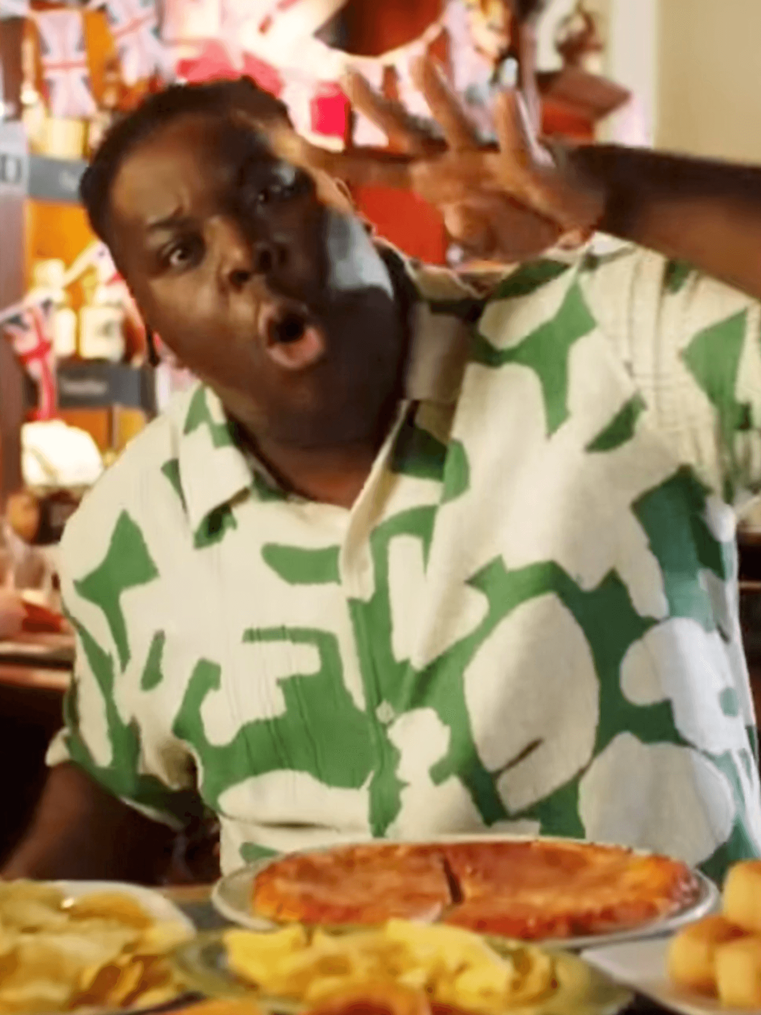 Hammed Animashaun, a Black man with short black hair and a green and white shirt, holds up four fingers while standing behind four plates of food in a pub.