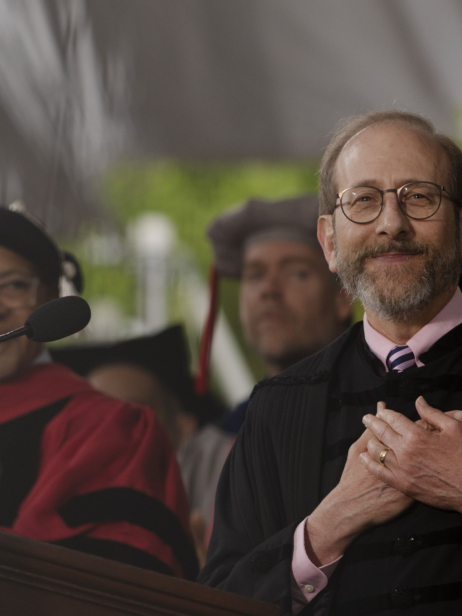 Harvard president Alan Garber, a bearded white man with rounded glasses, places both hands on his heart and smiles as he's applauded at the university's commencement ceremony.